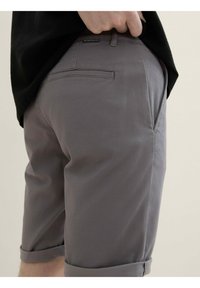 Pantaloni scurți gri din bumbac, cu manșete răsucite, confecționați dintr-un material neted, având un buzunar la spate și o etichetă mică a mărcii pe talie.