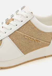 Balti odiniai sportbačiai su smėlio spalvos tekstūruotu audiniu, auksiniu "Michael Kors" logotipu ir baltais raišteliais. Turi lanksčią rusvą gumos padą.