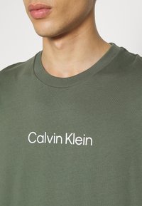 Oliwkowa bawełniana koszulka z okrągłym dekoltem i białym nadrukiem z logo "Calvin Klein" na przodzie. Gładka tekstura i luźny krój.