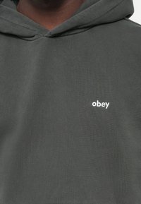 Hættetrøje i mørkegrå stof med en blød tekstur. Har et lille hvidt "obey" logo på brystet. Minimalistisk design.