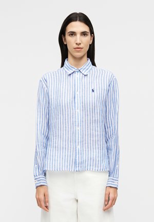 Frau trägt ein blau-weiß vertikal gestreiftes Hemd mit Knöpfen und kleinem gesticktem Logo, kombiniert mit weißen Hosen, steht vor einem schlichten Hintergrund.