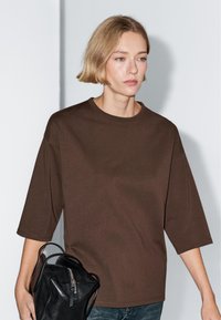 Young person with short blonde hair wears a brown oversized T-shirt and holds a black leather handbag against a plain gray background.