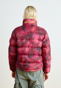Chaqueta acolchada roja y negra con un patrón de cuadros, cuello alto y logotipo amarillo de la marca en la parte posterior. La tela es brillante y acolchada.
