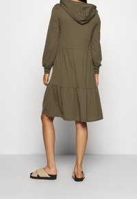 Olijfgroene hoodie jurk met lange mouwen, gelaagd ontwerp en zachte textuur, gecombineerd met lichtbeige instapsandalen.