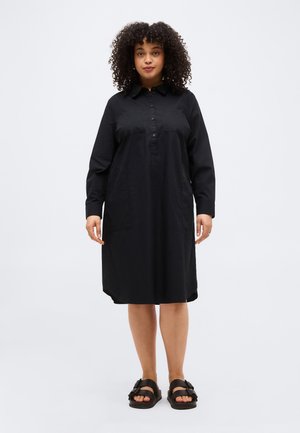 Kvinde med krøllet hår iført en langærmet sort kjole og sorte sandaler, der står foran en ensfarvet lys baggrund.