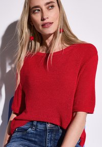 Roter Strickpullover mit lockerer Passform und weitem Halsausschnitt, kurzen Ärmeln, strukturiertem Muster, kombiniert mit dunkelblauen Jeans.