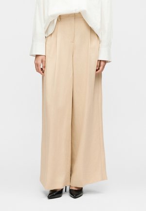 Bézs színű, bő szárú nadrág, fekete hegyes orrú magas sarkú cipővel és fehér hosszú ujjú inggel, egyszínű háttér előtt.