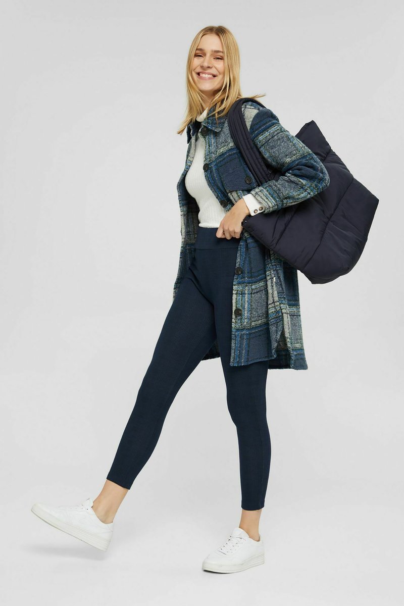 Cappotto a quadri blu e verde, maglione bianco a coste, leggings blu marino e sneakers bianche. La modella porta una borsa a tracolla nera e imbottita.