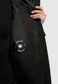 Sort jakke inderside med blød tekstur, logomærke med et ahornblad og en lynlåslomme. Indersidens stof er glat og mat.