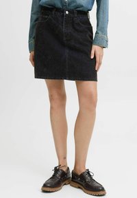 Kvinna klädd i en mörk denimkjol, blå denimjacka och bruna snörskor med tjocka sulor mot vit bakgrund.