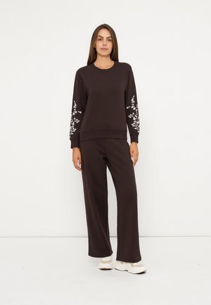 Donna che indossa un top a maniche lunghe marrone scuro con stampa floreale bianca sulle maniche e pantaloni a gamba larga abbinati, in piedi su uno sfondo bianco uniforme.