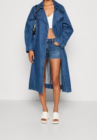 Osoba v dlouhém modrém džínovém kabátu, bílém crop topu, modrých džínových šortkách, bílých otevřených platformových podpatcích a s černou kabelkou přes rameno.