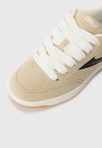 Beige semsket sko med hvite snører, sorte detaljer, og gummisåle. Har perforeringer og logo detaljer på hælen.