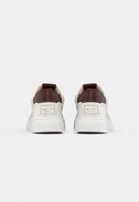 Adidași albi cu accente de piele maro, panouri laterale texturate și o talpă din cauciuc alb, având un logo și un tab în formă rotundă la călcâi.