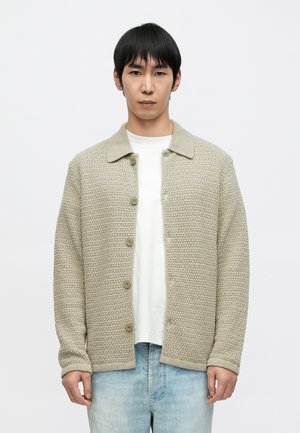 Beżowy dziergany sweter rozpinany z kołnierzem, z przodu zapinany na guziki, wykonany z fakturowanej tkaniny. Noszony na białej koszulce i jasnoniebieskich dżinsach.