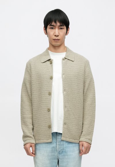 Beige knitted cardigan with a collar, featuring a button-up front and textured fabric. Worn over a white T-shirt and light blue jeans.