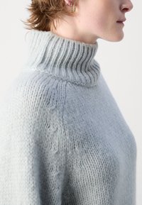 Světle šedý pletený rolák s žebrovaným límcem, který se vyznačuje měkkým, texturovaným povrchem a jemným kabelovým pletením na ramenou.