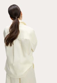 Chemise oversized jaune clair en tissu doux, avec un col rabattu et des épaules tombantes, portée avec un pantalon beige ample.