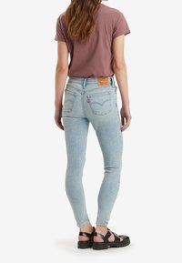 Jeans de talle alto de color azul claro con un diseño clásico, que cuentan con bolsillos traseros y una etiqueta marrón. Combinados con una camiseta ajustada de color malva y sandalias negras.