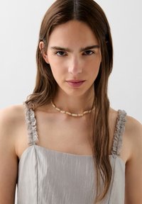 Abito in satin grigio con spalline arricciate, caratterizzato da una texture liscia. Il modello indossa una collana di perle di colore chiaro. Design semplice ed elegante.