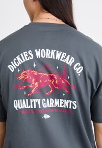 Pelēks kokvilnas T-krekls ar sarkanu un dzeltenu lapsu grafiku, tekstu "Dickies Workwear Co." un dekoratīviem elementiem baltā un tumši sarkanā krāsā.