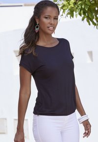 Zwart T-shirt met korte mouwen en een ronde hals, gecombineerd met witte jeans en meerdere witte armbanden, tegen een lichte achtergrond.