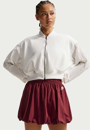 Femme aux cheveux tressés portant une veste blanche zippée et un short de sport Nike bordeaux avec une taille élastique, debout les mains sur les hanches.