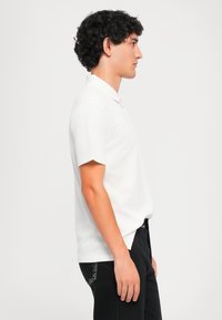 Νεαρός άντρας με σγουρά μαλλιά, φορώντας λευκό polo μπλουζάκι κοντό μανίκι και μαύρα τζιν, που στέκεται σε προφίλ μπροστά από απλή επιφάνεια.