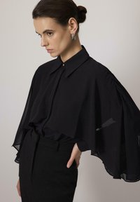 Black oversized blouse with a collar and sheer, flowing sleeves. Smooth fabric, featuring a row of buttons and a fitted waistband.