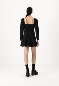 Vestido negro con mangas largas y espalda cuadrada, con un cuerpo fruncido. La falda se expande, combinado con zapatos de plataforma negros y calcetines.