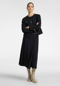 Giacca nera su misura con accessori argentati, abbinata a una lunga gonna nera. Stivali al ginocchio beige completano l'outfit.