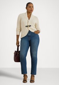 Moteris, vilkinti kreminės spalvos pintos tekstūros kardiganą su sagtimi, mėlynais džinsais, rudos spalvos aukštakulniais sandalais, laikanti tamsiai raudoną rankinę, stovinti priešais paprastą foną.