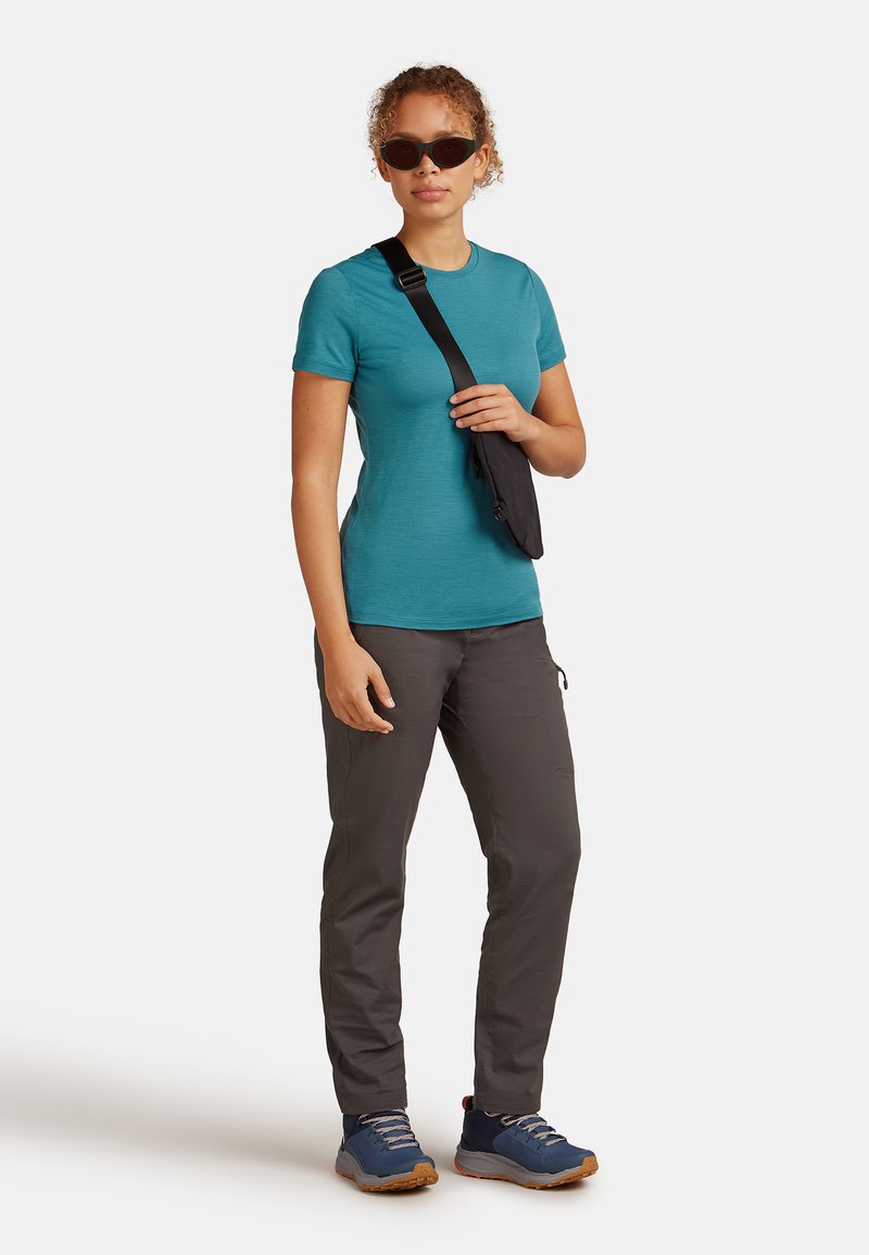 Teal T-shirt met korte mouwen, grijze broek en blauwe wandelschoenen. Zwarte schoudertas. Krullend haar en zonnebril. Buitenkleding met functioneel ontwerp.