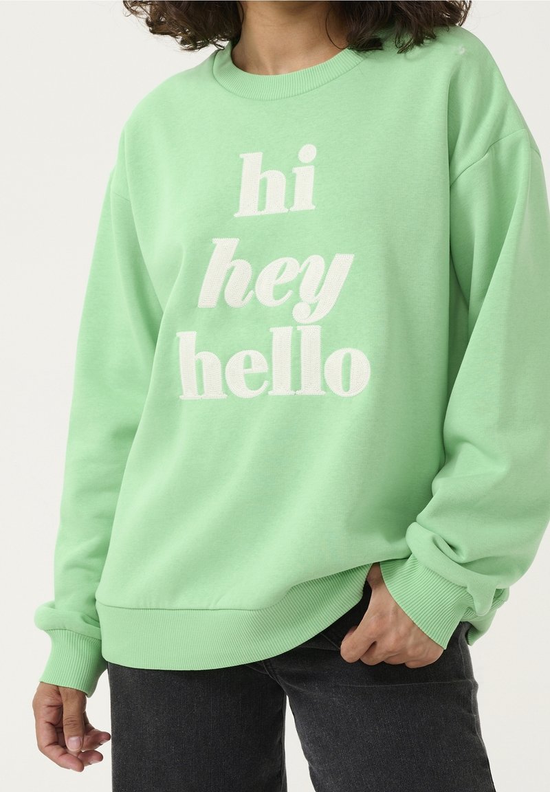 Personne vêtue d'un sweat-shirt vert clair avec un texte blanc disant "hi hey hello", debout avec une main dans la poche d'un jean noir.