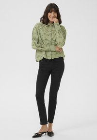 Chemise boutonnée vert clair avec des motifs en œillets, ourlet brodé, associée à un jean slim noir et des escarpins à bout pointu noirs.