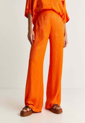 Person iført lyse orange vidde bukser med subtile geometriske mønstre og brune platåsandaler mod en ensfarvet baggrund.
