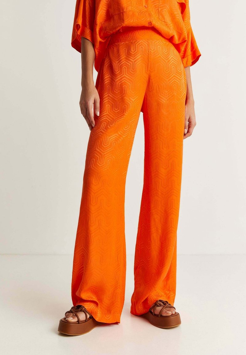 Personne portant un pantalon large orange vif avec des motifs géométriques subtils et des sandales à plateforme marron sur fond uni.