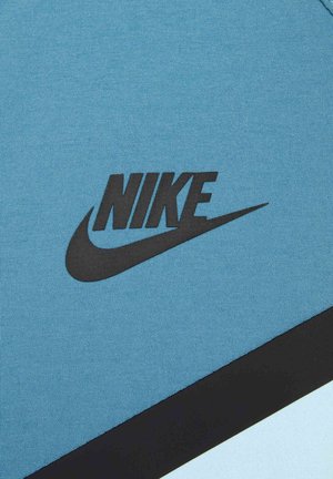 Nike urheiluasu vaaleansinisestä kankaasta, jossa on musta logo ja muotoiluelementtejä. Tekstuurin pinta on sileä, ja siihen on lisätty kontrastina musta saumaviiva.