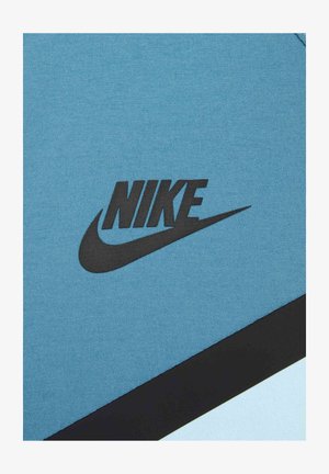 Nike urheiluasu vaaleansinisestä kankaasta, jossa on musta logo ja muotoiluelementtejä. Tekstuurin pinta on sileä, ja siihen on lisätty kontrastina musta saumaviiva.