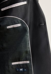 Veste de costume noire partiellement ouverte montrant une doublure intérieure verte foncée à motif avec bordure blanche et une étiquette indiquant Nova Fides Italy.