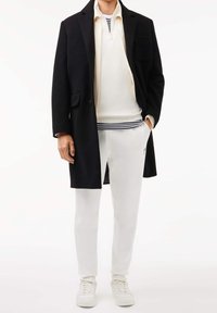 Černý kabát přes bílou košili na zip s tmavě modrými pruhy, v kombinaci s bílými kalhotami a bílými teniskami. Má úzký střih a hladké textury.