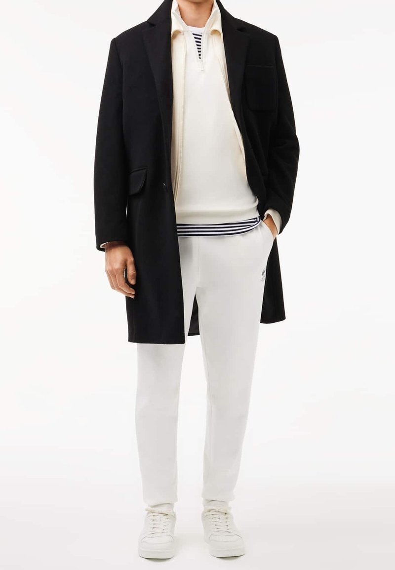 Černý kabát přes bílou košili na zip s tmavě modrými pruhy, v kombinaci s bílými kalhotami a bílými teniskami. Má úzký střih a hladké textury.