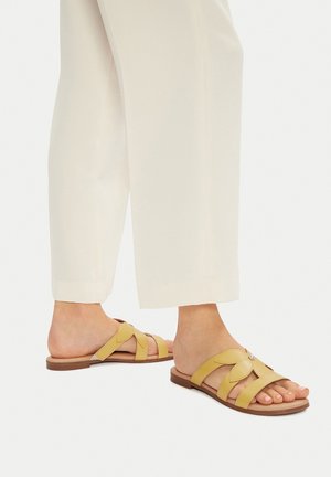 Sandalias de cuero amarillo con diseño cruzado, que presentan un detalle decorativo. La suela es marrón y plana, adecuada para uso casual.