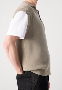 Beige gestrickte Weste mit V-Ausschnitt und kurzen gerippten Ärmeln, getragen über einem weißen Hemd. Die Weste hat einen lockeren Schnitt und eine glatte Textur.