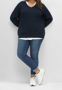 Kobieta ubrana w granatowy sweter w warkocze, białą koszulę, niebieskie jeansy podwinięte na nogawkach i białe trampki, stoi z nogami skrzyżowanymi na jednolitym tle.