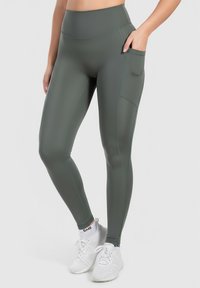 Zelené legíny s vysokým pasem z elastick�ého matného materiálu, se boční kapsou a kontrastním šitím. Nosené s bílými teniskami.