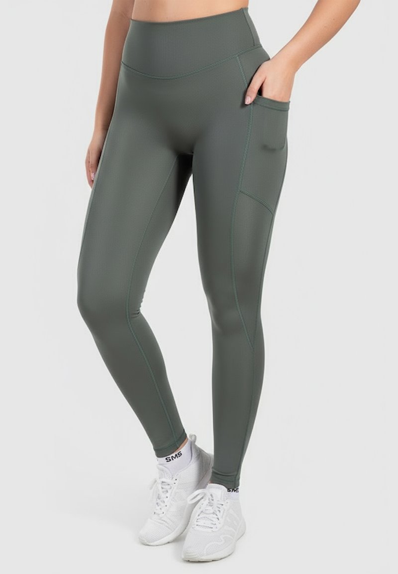 Zelené legíny s vysokým pasem z elastického matného materiálu, se boční kapsou a kontrastním šitím. Nosené s bílými teniskami.
