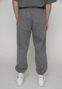 Graue Jogginghose mit elastischem Bund und gerippten Bündchen, mit einer Gesäßtasche. Getragen mit weißen Sneakern vor neutralem Hintergrund.