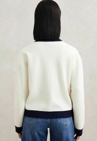 Bela pulover s mornarsko modrim rebrastim ovratnikom in robom, ki ima mehak vzorec. Uporabljen z modrimi kavbojkami, kar prikazuje sproščen zadnji pogled.