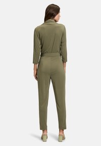 Tuta verde oliva con maniche lunghe, vita aderente e gambe a sigaretta. Realizzata in tessuto morbido, presenta un colletto e dettagli minimi.